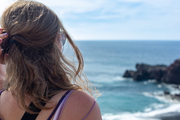 Giovane donna con lunghi capelli biondi seduti sulla spiaggia e guardando l'orizzonte. Oceano blu e costa rocciosa.