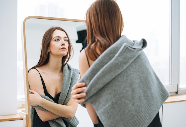 Giovane donna con lunghi capelli scuri in abito nero elegante da sera con schiena aperta vicino allo specchio all'interno bianco