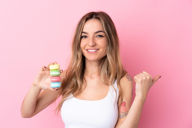 Giovane donna con macarons sopra isolato muro rosa che punta verso il lato per presentare un prodotto