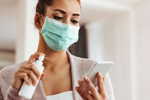 Giovane donna con maschera facciale che disinfetta il suo smartphone a casa