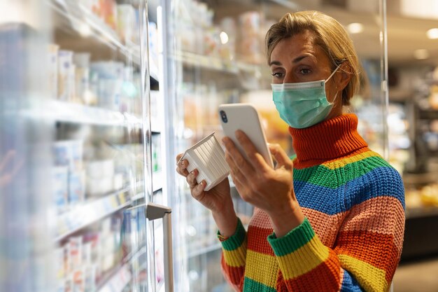 Giovane donna con maschera facciale che utilizza il telefono cellulare e acquista generi alimentari nella sezione congelatore del supermercato durante la pandemia di Coronavirus