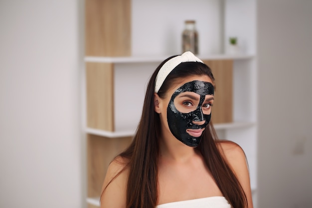 Giovane donna con maschera nera detox al carbonio sul viso, ragazza adolescente si prende cura della pelle grassa