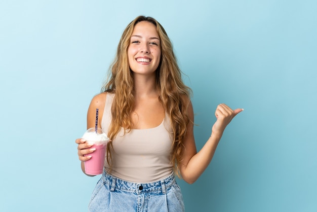 Giovane donna con milkshake alla fragola isolato sulla parete blu che punta di lato per presentare un prodotto