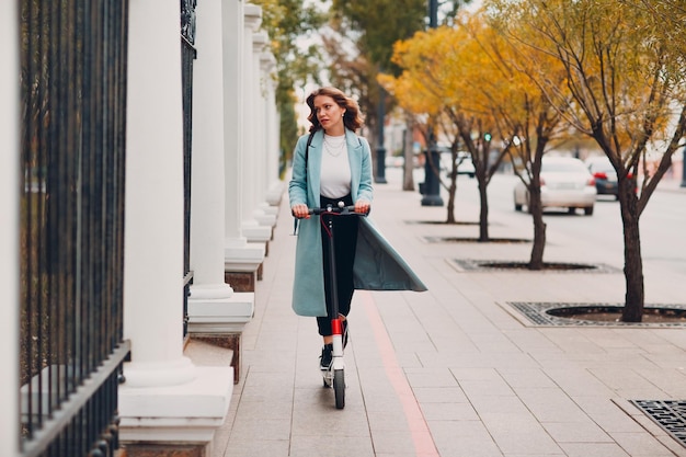 Giovane donna con scooter elettrico in cappotto blu in città