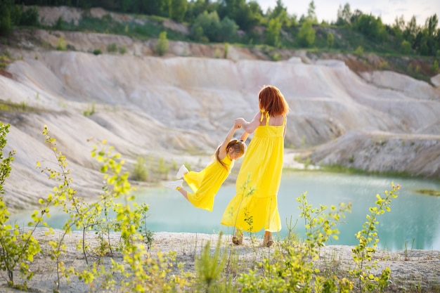 Giovane donna con sua figlia in un vestito giallo vicino al lago con acqua azzurra e alberi verdi. Concetto di relazione familiare felice