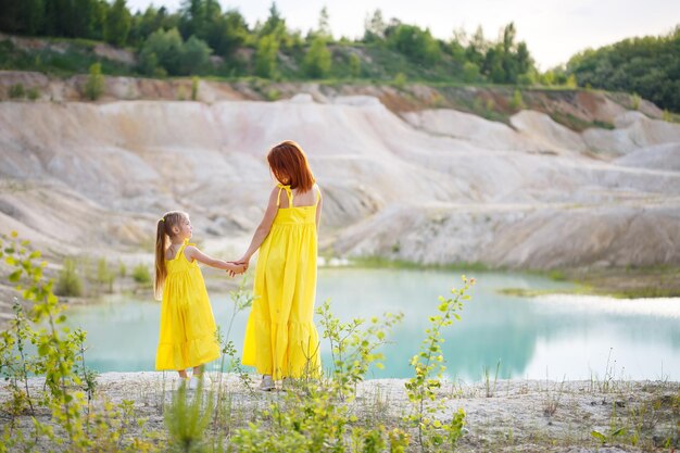 Giovane donna con sua figlia in un vestito giallo vicino al lago con acqua azzurra e alberi verdi. Concetto di relazione familiare felice