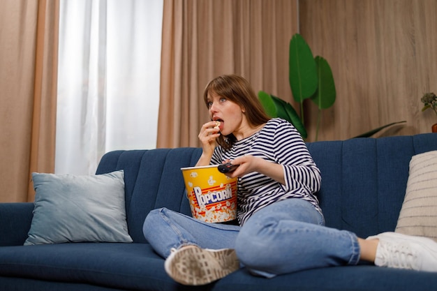 Giovane donna con telecomando che guarda la tv e mangia popcorn sul divano