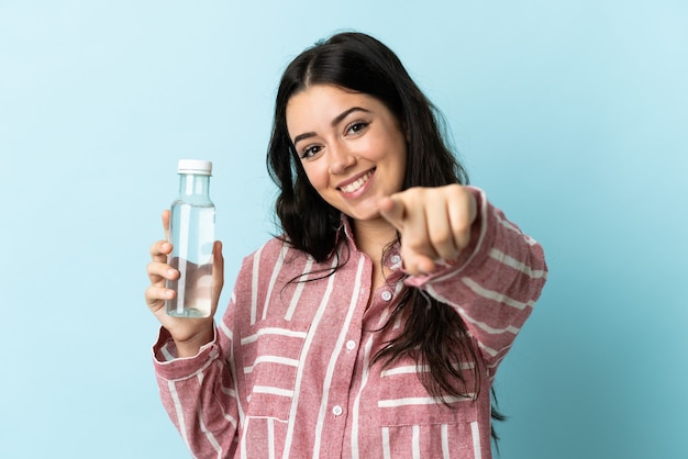 Giovane donna con un'acqua isolata sul muro blu punta il dito contro di te con un'espressione sicura