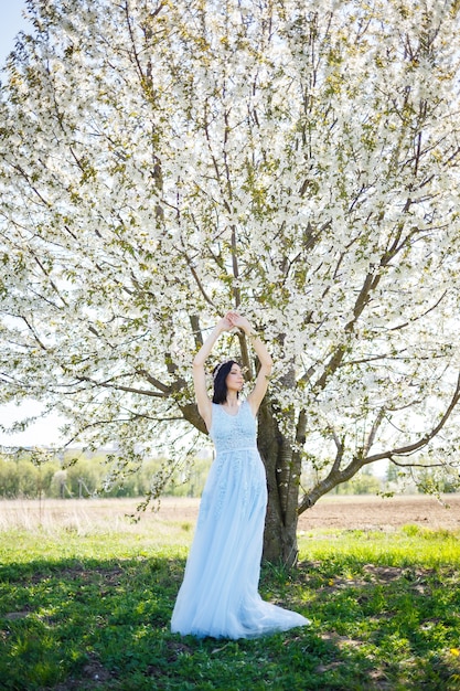 Giovane donna con un bel trucco con un cerchio vintage in un vestito blu chiaro con un bouquet in posa su uno sfondo di fogliame verde in un parco. Ragazza attraente del ritratto fresco con i fiori bianchi.