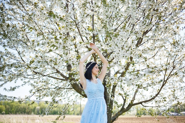 Giovane donna con un bel trucco con un cerchio vintage in un vestito blu chiaro con un bouquet in posa su uno sfondo di fogliame verde in un parco. Ragazza attraente del ritratto fresco con i fiori bianchi.