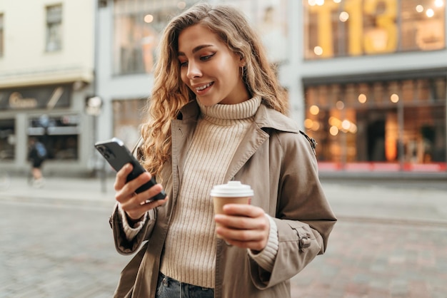 Giovane donna con un cappotto d'autunno che cammina con uno smartphone e una tazza di caffè in una strada della città