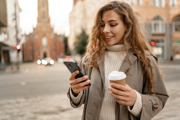 Giovane donna con un cappotto d'autunno che cammina con uno smartphone e una tazza di caffè in una strada della città