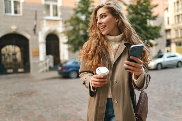 Giovane donna con un cappotto d'autunno che cammina con uno smartphone e una tazza di caffè in una strada della città
