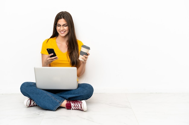 Giovane donna con un computer portatile che si siede sul pavimento che tiene il caffè da portare via e un cellulare