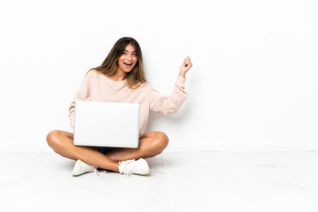 Giovane donna con un computer portatile che si siede sul pavimento isolato su bianco che fa il gesto della chitarra