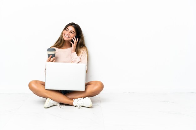 Giovane donna con un computer portatile che si siede sul pavimento isolato sul muro bianco che tiene il caffè da portare via e un cellulare