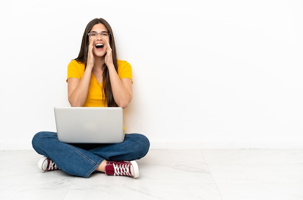 Giovane donna con un computer portatile seduta sul pavimento che grida e annuncia qualcosa