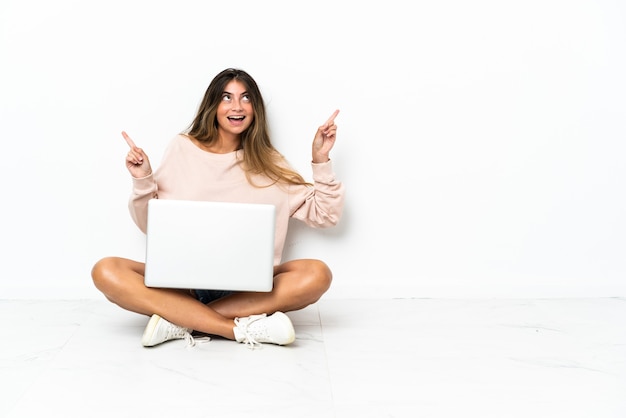 Giovane donna con un computer portatile seduto sul pavimento isolato sul muro bianco che punta il dito ai laterali e felice