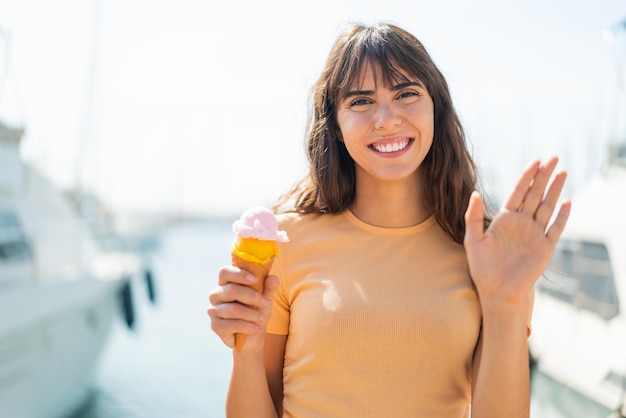 Giovane donna con un gelato alla cornetta all'aperto che saluta con la mano con un'espressione felice