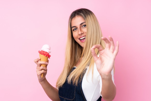 Giovane donna con un gelato di cornetta sopra la parete rosa isolata che mostra segno giusto con le dita