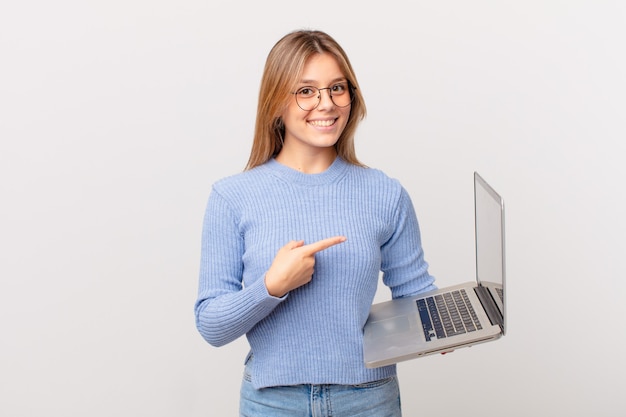 Giovane donna con un laptop che sorride allegramente, si sente felice e indica il lato