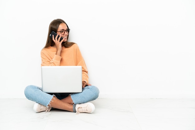 Giovane donna con un laptop seduta sul pavimento che tiene una conversazione con il telefono cellulare con qualcuno