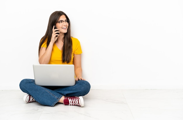 Giovane donna con un laptop seduta sul pavimento che tiene una conversazione con il telefono cellulare con qualcuno