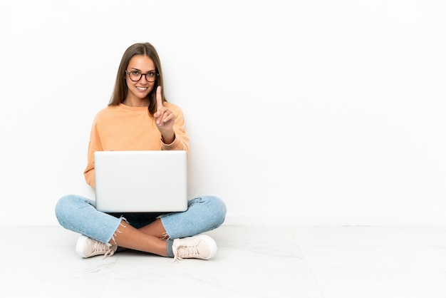 Giovane donna con un laptop seduto sul pavimento che mostra e alza un dito