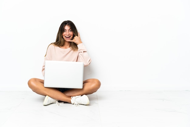 Giovane donna con un laptop seduto sul pavimento isolato su sfondo bianco che fa il gesto del telefono. Richiamami segno