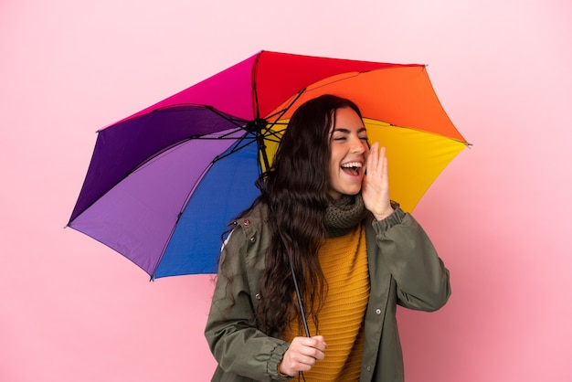 Giovane donna con un ombrello isolato su sfondo rosa che grida con la bocca spalancata di lato