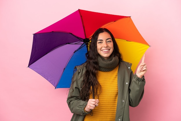 Giovane donna con un ombrello isolato su sfondo rosa che mostra e alzando un dito in segno del meglio