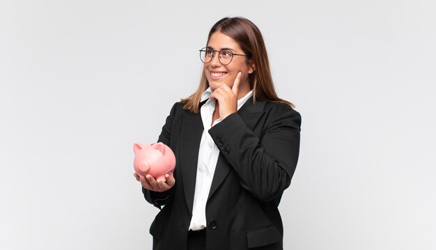 Giovane donna con un salvadanaio sorridente felicemente e fantasticando o dubitando, guardando al lato