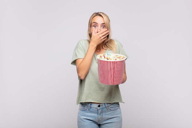 Giovane donna con un secchio di pop corn che copre la bocca con le mani con un'espressione scioccata e sorpresa, mantenendo un segreto o dicendo oops