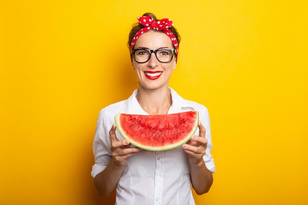 Giovane donna con un sorriso in una fascia rossa e occhiali tiene un'anguria su una parete gialla.