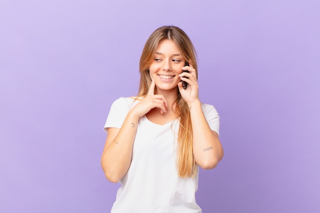 Giovane donna con un telefono cellulare che sorride con un'espressione felice e sicura con la mano sul mento