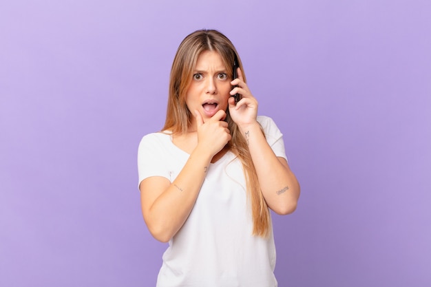 Giovane donna con un telefono cellulare con la bocca e gli occhi spalancati e la mano sul mento