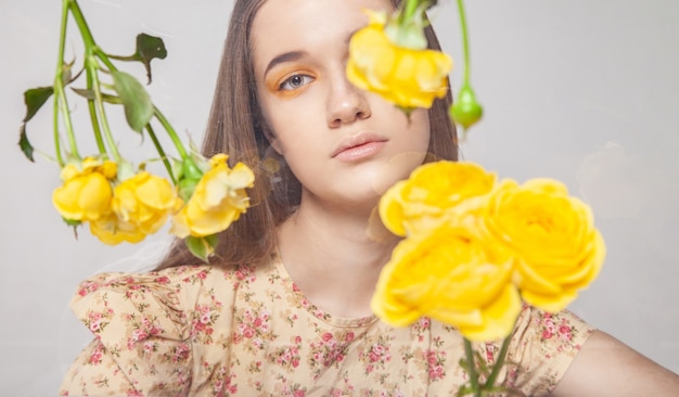 Giovane donna con un trucco elegante che guarda la telecamera attraverso un contenitore di vetro con fiori gialli stabilizzati su sfondo grigio