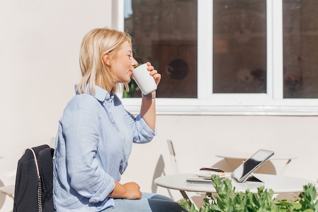 Giovane donna con una camicia blu che lavora su un notebook in un bar e beve caffè