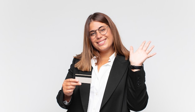 Giovane donna con una carta di credito che sorride allegramente e allegramente, agitando la mano, dandoti il benvenuto e salutandoti o salutandoti