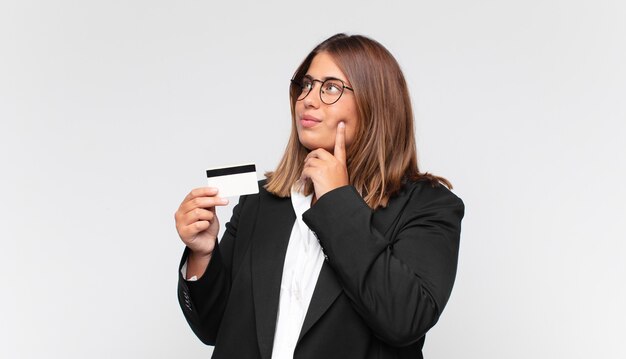 giovane donna con una carta di credito sorridente felicemente e fantasticando o dubitando, guardando al lato