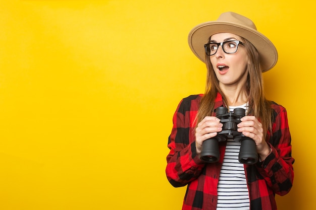 Giovane donna con una faccia sorpresa in un cappello e una camicia a quadri che tiene il binocolo nelle sue mani su uno sfondo giallo.