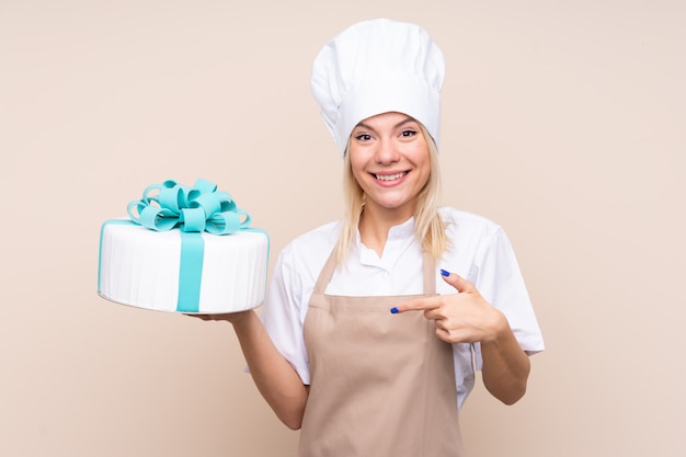 Giovane donna con una grande torta sopra la parete isolata con espressione facciale a sorpresa