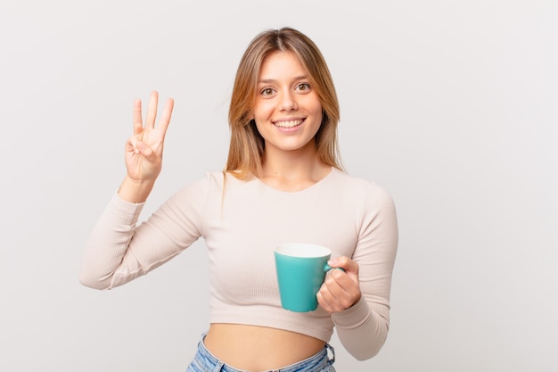 giovane donna con una tazza da caffè sorridente e dall'aspetto amichevole, mostrando il numero tre