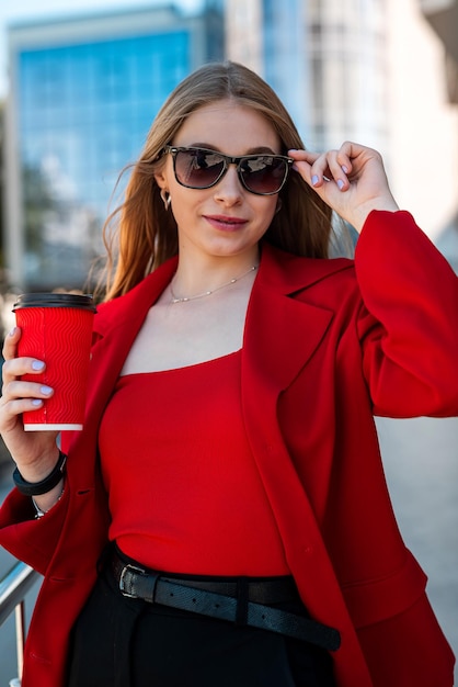 Giovane donna con una tazza di caffè in mano con il trucco e gli occhiali che guarda di lato mentre è in piedi vicino al suo ufficio Ritratto di una ragazza d'affari vicino a un edificio di vetro