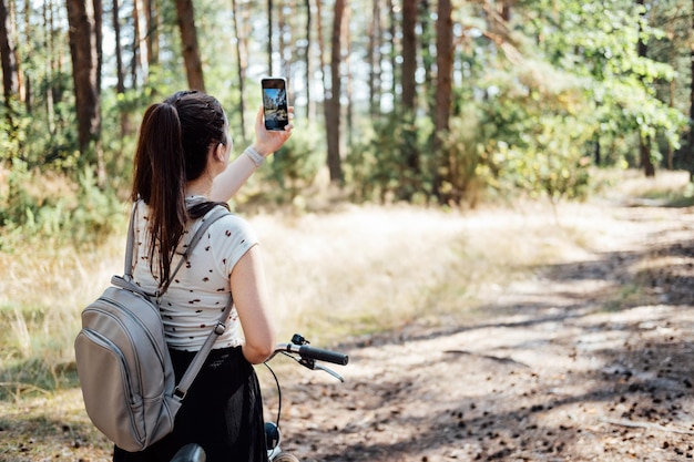Giovane donna con zaino in bicicletta e scattare foto sul cellulare su sfondo foresta di pini ragazza