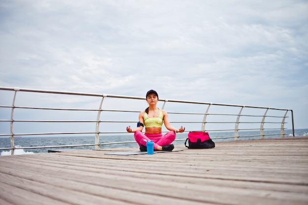 Giovane donna concentrata che si siede su una stuoia di yoga nella posa del loto sulla spiaggia