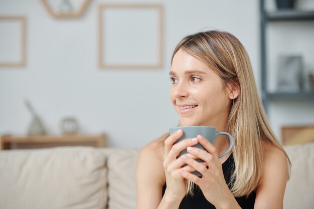 Giovane donna contemporanea con un sorriso a trentadue denti e lunghi capelli biondi che tiene in mano una tazza con una bevanda calda mentre si gode il caffè mattutino