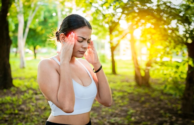 Giovane donna corridore che si massaggia la testa con l'emicrania in un parco Donna corridore con mal di testa e fatica nel parco Donna corridore con mal di testa in un parco Atleta ragazza con emicrania in un parco