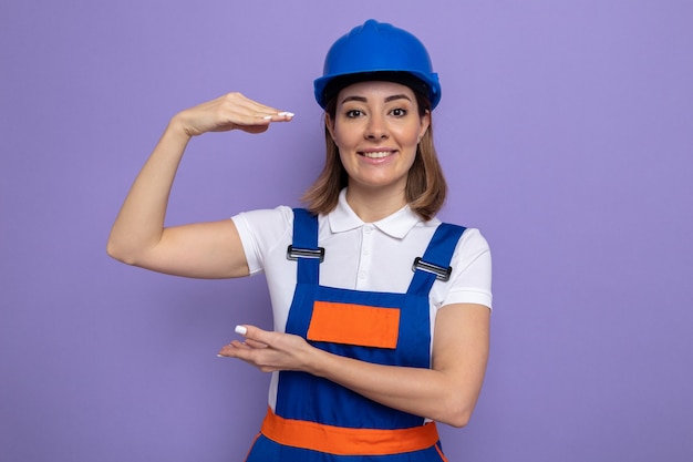 Giovane donna costruttore in uniforme da costruzione e casco di sicurezza sorridente fiducioso che mostra gesto di dimensioni con le mani in piedi sul muro viola