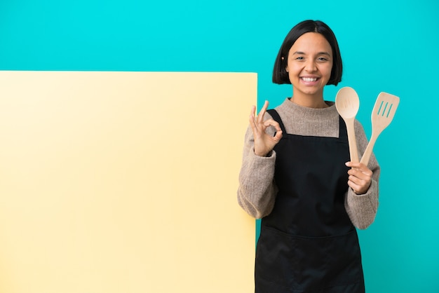 Giovane donna cuoca di razza mista con un grande cartello isolato su sfondo blu che mostra segno ok con due mani